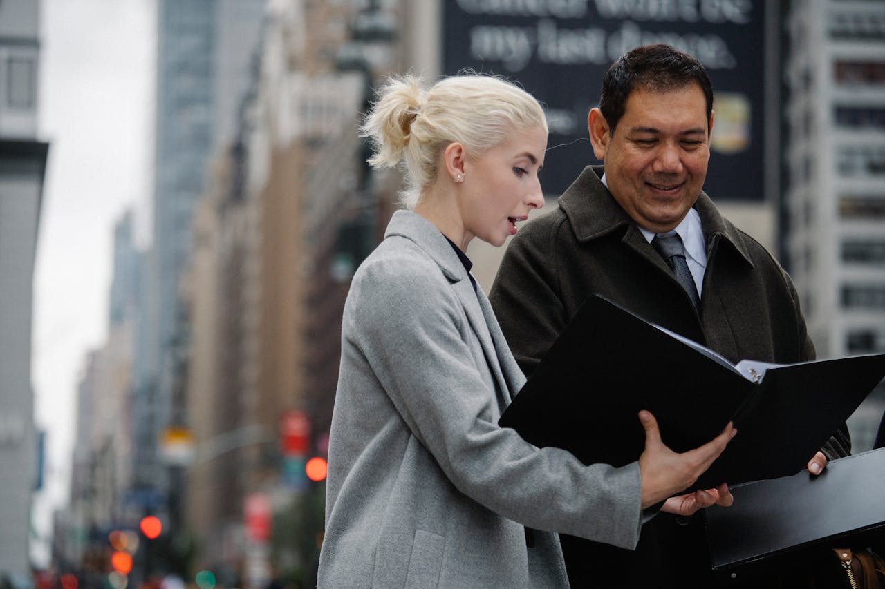 Two professionals conversing and examining documents in an urban outdoor setting.