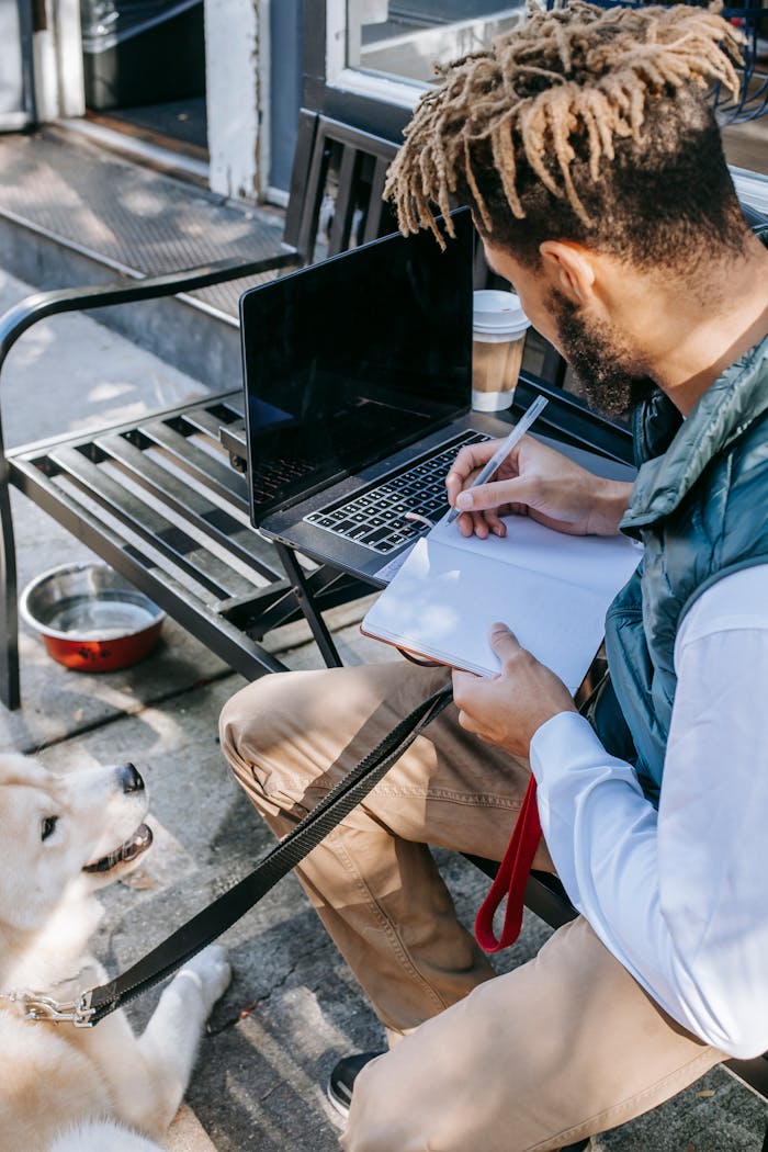 Man taking notes on notepad with a dog by his side; working remotely in an urban setting.